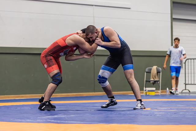 Two male wrestlers in intense clinch, red vs blue singlet, both leaning in aggressively on an orange and blue mat.