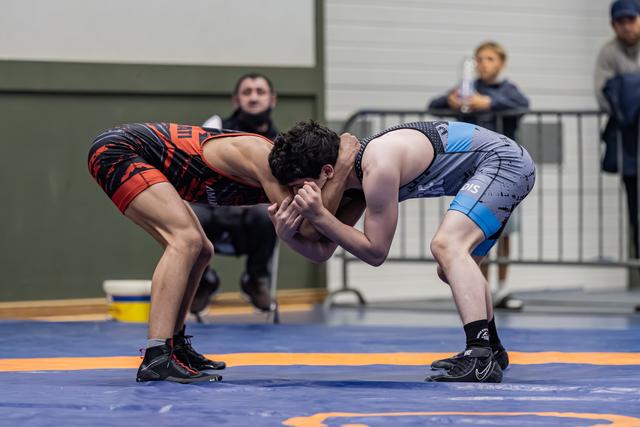 Two wrestlers locked in an intense clinch, bent low in a defensive stance on a blue competition mat.