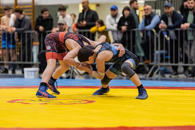 Two young wrestlers lock in an intense clinch on a yellow mat, crowd watching closely behind metal barriers.