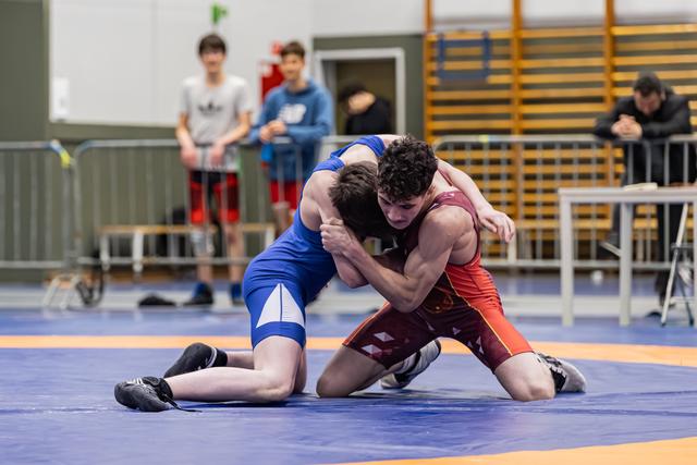 Two young wrestlers locked in an intense clinch on the blue mat, both straining with focused determination during a match.