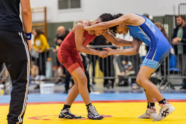 Two young female wrestlers in red and blue singlets lock hands in an intense stance on a yellow competition mat.