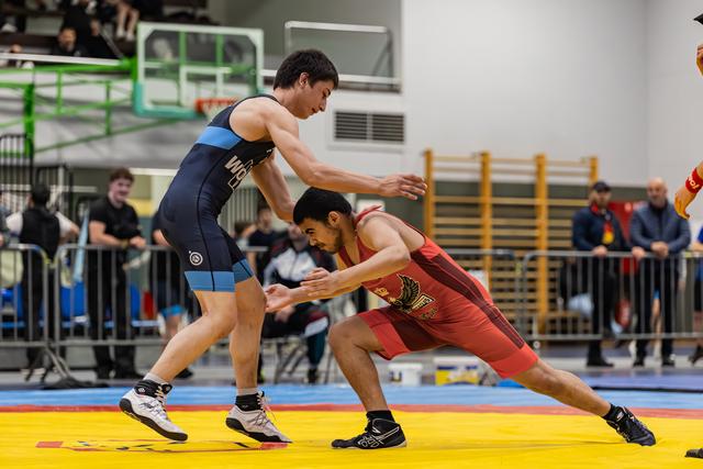 Two young wrestlers engage in a tense standoff on a yellow mat, one in navy blue, one in red, both crouching low.