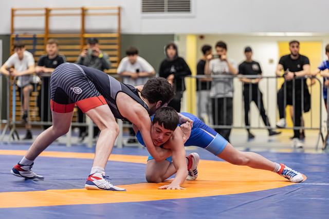 Young wrestler in blue singlet fights to escape a takedown attempt on an orange mat, crowd watching behind barriers.
