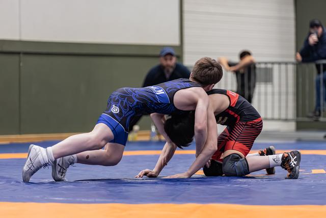 Young wrestler in blue singlet drives forward aggressively while opponent in black and red struggles on the mat.