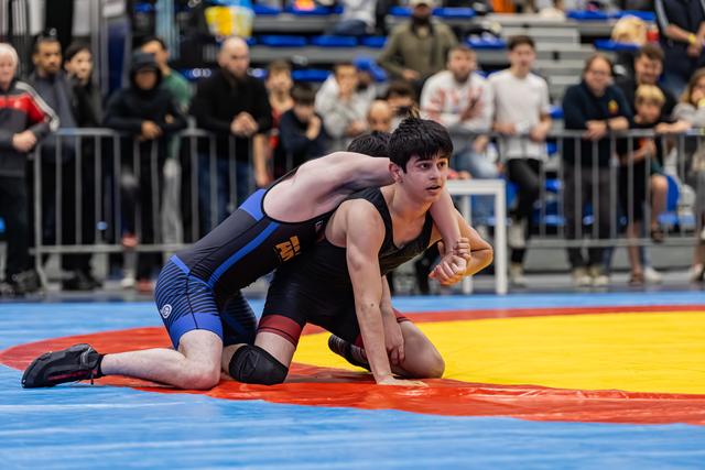 Young wrestler in black singlet holds position on mat, looking focused, as opponent in blue applies pressure from behind.