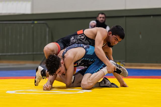 Wrestler in black tiger singlet dominates opponent on yellow mat, both focused and intense during indoor bout.