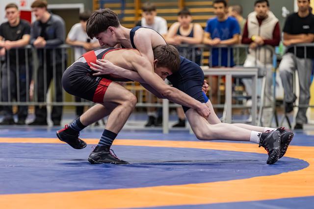 Two young male wrestlers locked in a takedown struggle on a blue and orange mat, spectators watching intently behind barriers.