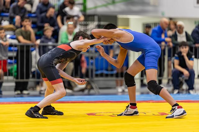 Two young wrestlers in black and blue singlets face off in a tense clinch on a yellow mat before a packed crowd.