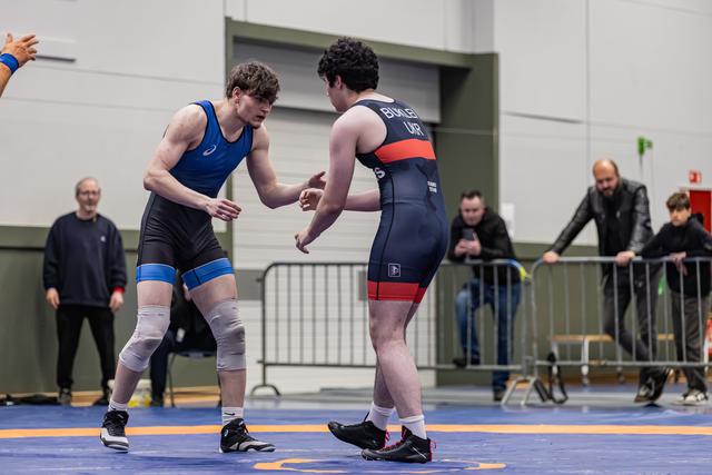 Two young male wrestlers reach toward each other in a pre-match handshake on a blue mat, spectators watching behind barriers.