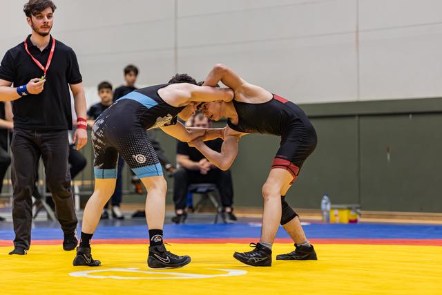 Two young wrestlers grapple head-to-head on a yellow mat, locked in a tense clinch, referee watching closely nearby.
