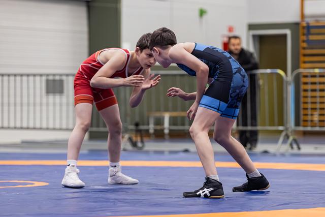 Two young wrestlers in red and blue singlets crouch in tense standoff, hands raised, on a competition mat indoors.