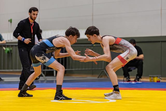 Two teen wrestlers crouch in tense standoff, hands reaching toward each other on a yellow mat, referee watching closely.