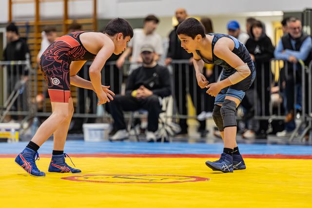 Two young wrestlers crouch in defensive stances, locked in intense eye contact before engaging on a yellow competition mat.