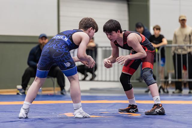 Two young wrestlers in blue and red singlets crouch in tense standoff on blue mat, spectators watching behind barriers.