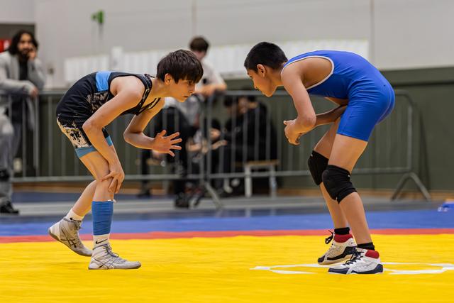 Two young wrestlers crouch in a tense standoff on a yellow mat, focused and ready to engage in a gym.