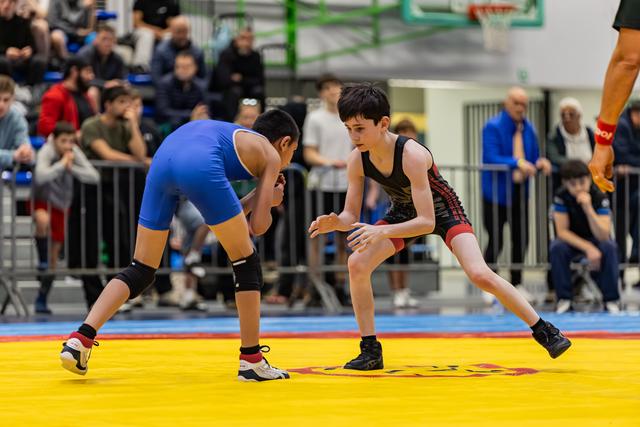 Two young wrestlers in blue and black singlets crouch in an intense standoff on a yellow competition mat, crowd watching.
