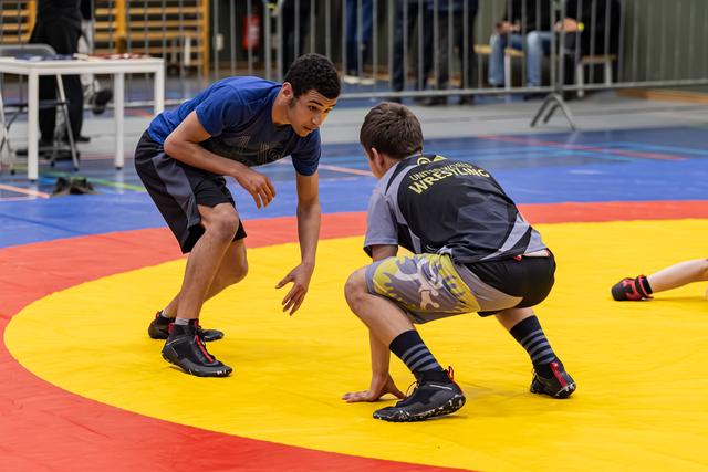 Two young wrestlers in crouched stances face each other intently on a colorful competition mat indoors.