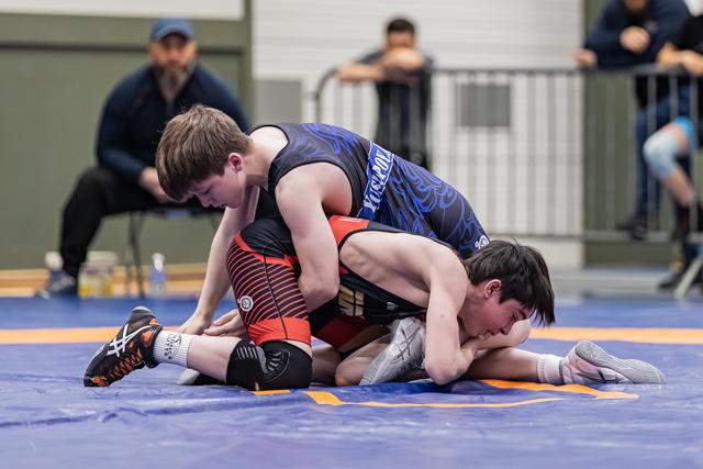 Young wrestler in blue singlet controls opponent on blue mat, spectators watching in background during indoor meet.