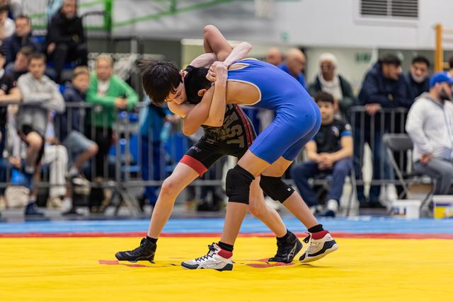 Two young wrestlers locked in a grappling hold on a yellow mat, crowds watching in a busy sports hall.