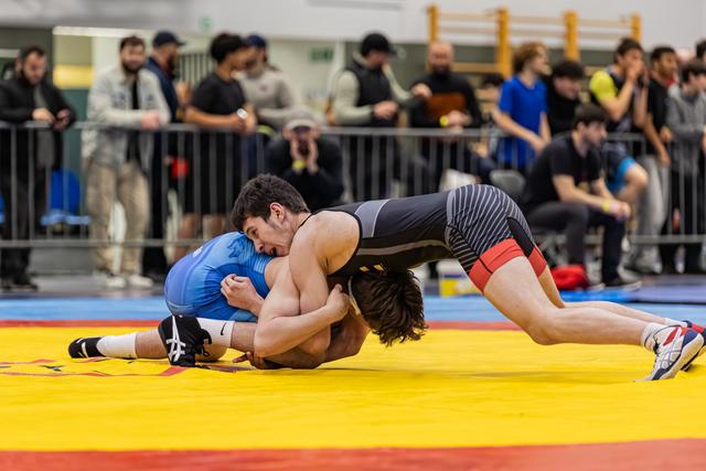Wrestler in black singlet aggressively drives blue-clad opponent toward the mat, crowd watching intently in background.