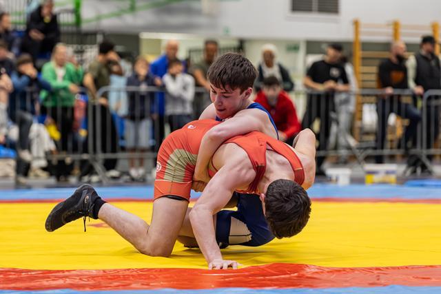 Young wrestler in blue dominates opponent in red, pinning him to the yellow mat during an indoor tournament.