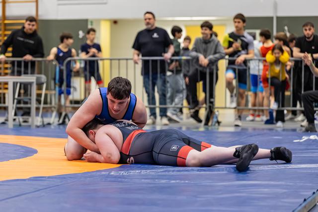 Young wrestler in blue singlet pins opponent face-down on mat as crowd watches intently in background.