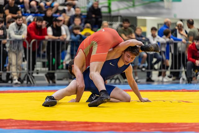 Wrestler in blue singlet braces against opponent in red attempting a takedown on a yellow competition mat.