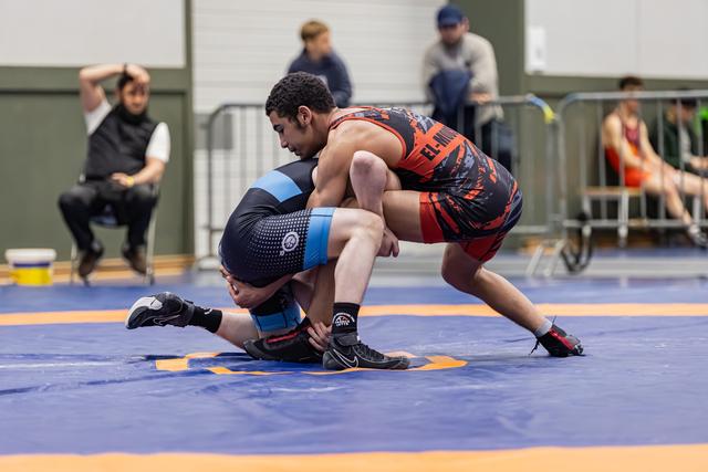 Young wrestler in red singlet drives opponent toward the mat in a powerful double-leg takedown during a match.