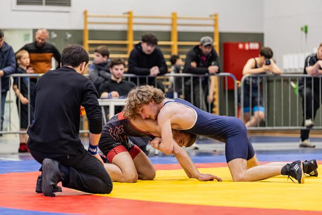 Young wrestler in navy singlet dominates opponent on mat while referee watches closely indoors.