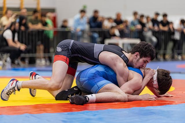 Wrestler in dark singlet pins opponent in blue to the red mat, crowd watching in background.