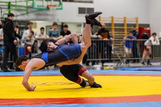 Young wrestler in blue singlet performs a powerful suplex throw, launching opponent airborne on colorful competition mat.