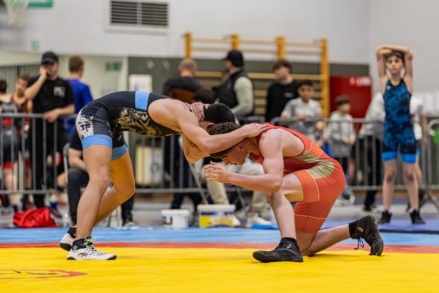 Two wrestlers battle on a yellow mat, one in red kneeling while being controlled in a headlock by opponent in black.