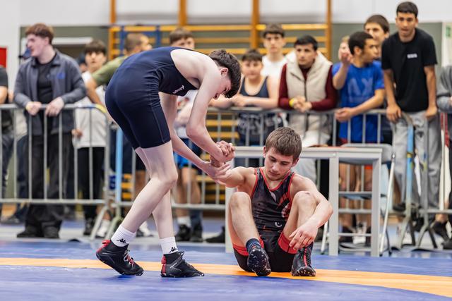 Blue singlet wrestler extends hand to help seated opponent rise from mat, crowd watches in background.