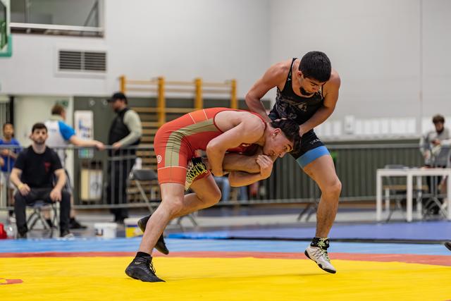 Wrestler in black singlet aggressively controls bent-over opponent in red during indoor match on yellow mat.