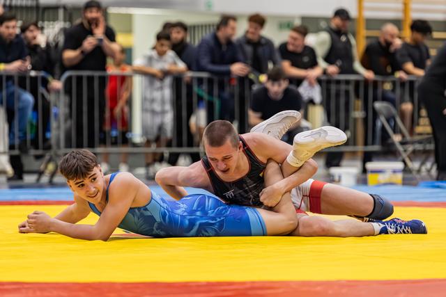 Young wrestler in blue singlet smiles while being pinned by opponent in black, on a yellow and red mat.