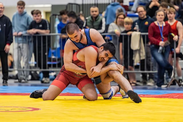 Wrestler in red singlet controls smiling opponent on yellow mat, crowd watching in background.