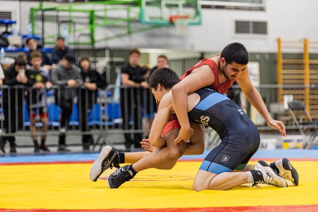 Wrestler in red singlet drives opponent down onto the yellow mat during an intense indoor match, crowd watching.