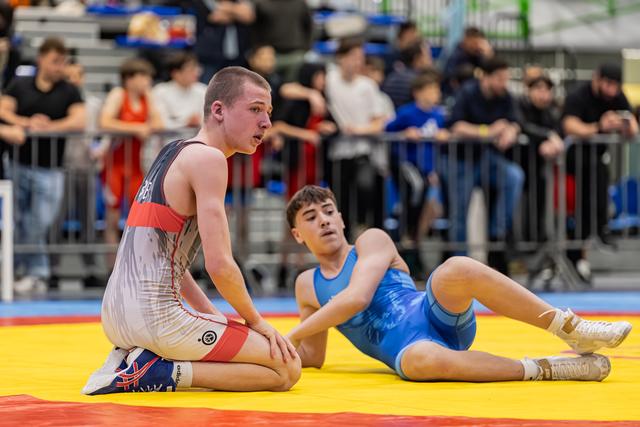 Young wrestler kneels confidently on yellow mat while opponent lies sprawled behind him, crowd watching in background.