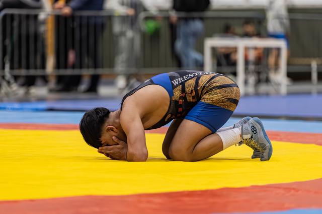 Young wrestler kneels on the mat, face buried in hands, expressing deep emotion after a match.
