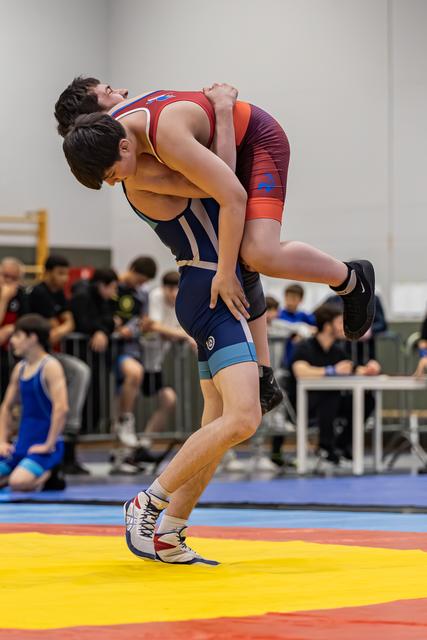 Young wrestler in navy singlet lifts opponent clean off the mat in a powerful body-lock during an indoor tournament.
