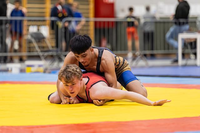 Young wrestler in red singlet pinned to the yellow mat, opponent in black applying pressure in an indoor arena.