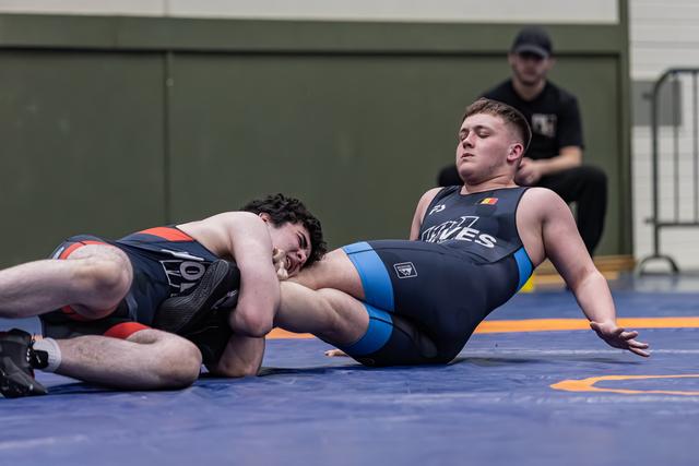 Young wrestler being pinned on his back while opponent maintains control, referee watching closely in background.