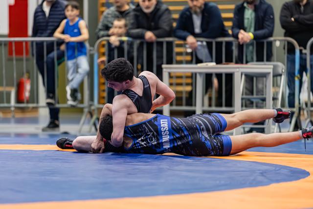 Athlete in black singlet pins opponent in blue 'Mussati' singlet to orange mat, crowd watching behind barriers.
