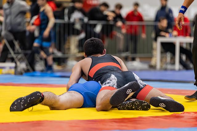 Wrestler in black singlet pins opponent in blue to the yellow mat during an indoor tournament match.