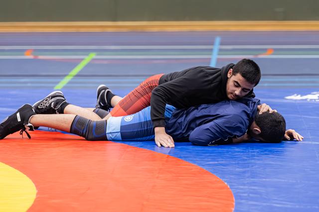 Young wrestler in black top pins opponent in blue singlet to the mat, maintaining dominant top position.
