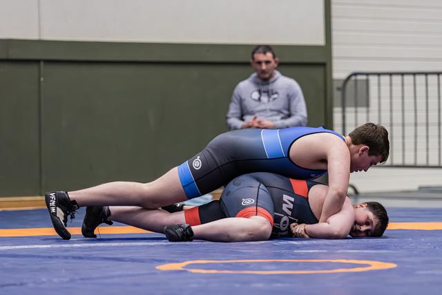 Young wrestler in blue singlet pins opponent to the mat while a coach watches intently from the background.
