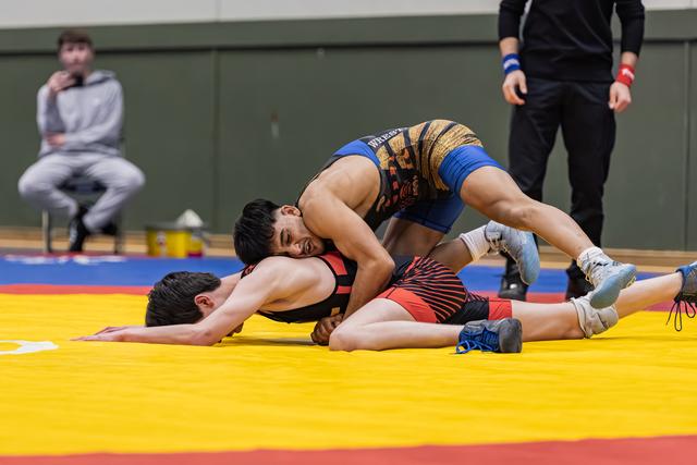 Blue-suited wrestler pins opponent face-down on yellow mat, referee watching nearby, spectator seated in background.