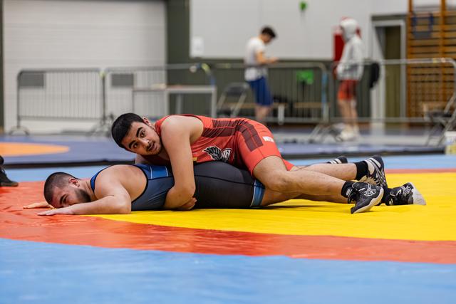 Young wrestler in red singlet pins opponent to the mat, maintaining control with focused expression in indoor sports hall.