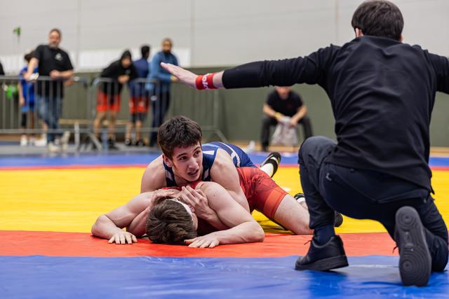 Young wrestler in blue pins opponent face-down on mat as referee kneels and raises hand to signal.
