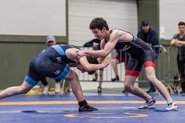 Young wrestler in dark singlet aggressively drives opponent's head down during intense match on blue mat.